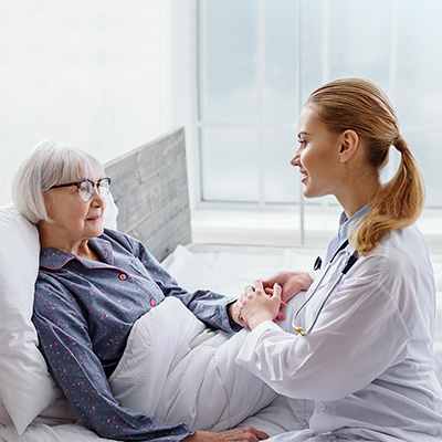 female nurse sitting on the bed of an elderly resident and holding hands